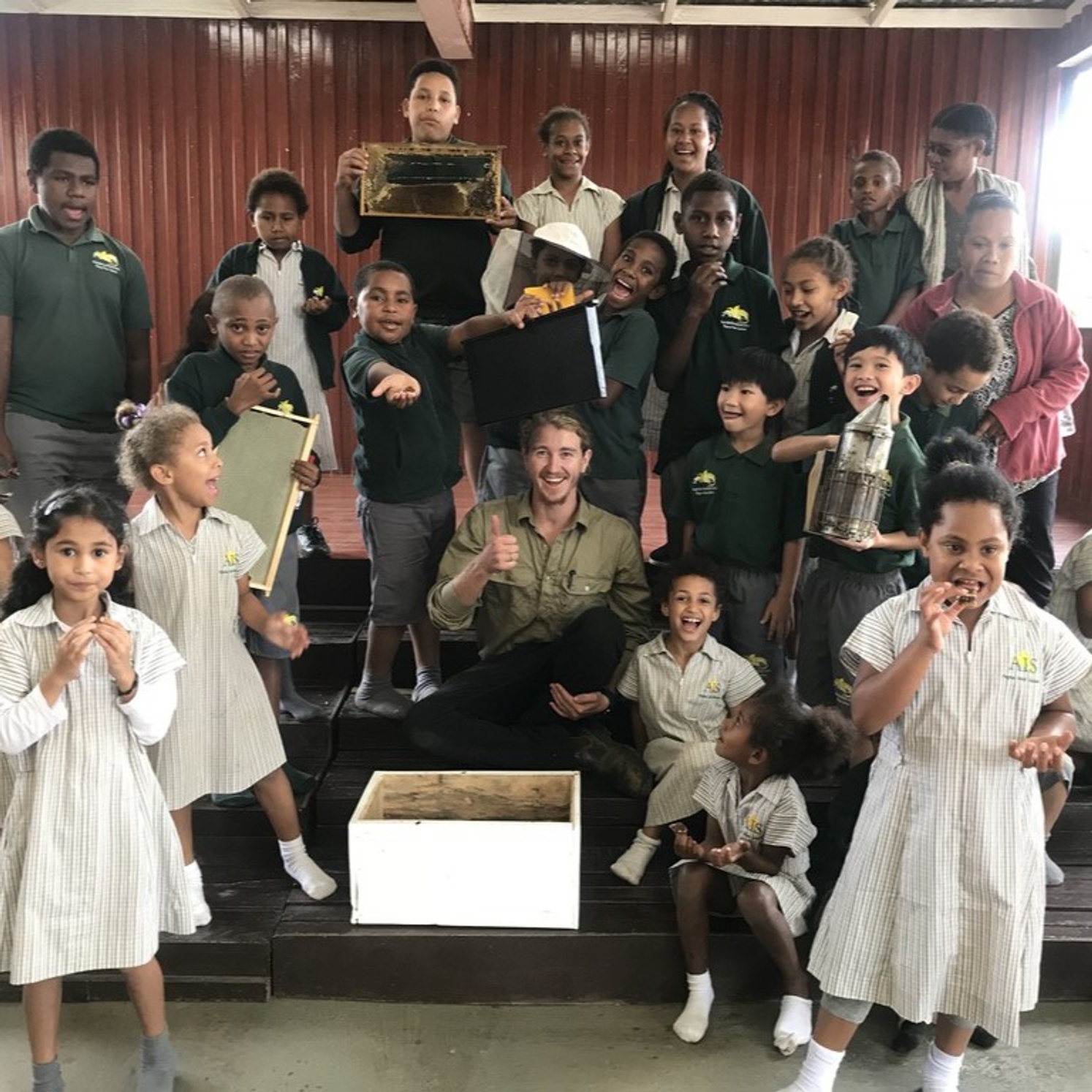 Man sitting, surrounded by happy smiling young children holding bee keeping implements