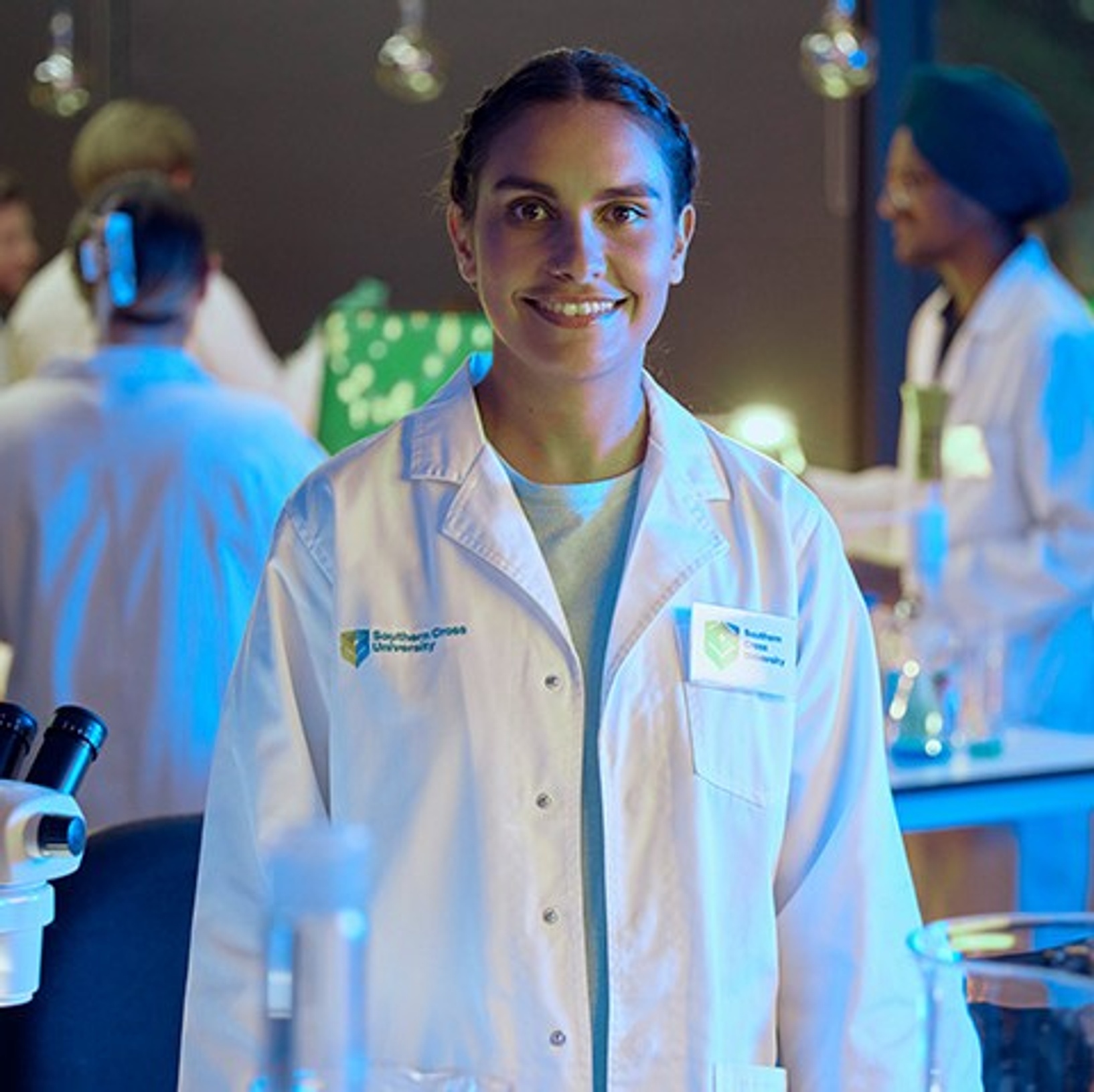 Ashlee Cable, a Bachelor of Science student, stands in a Southern Cross classroom laboratory with lab coat while her fellow students work behind her surrounded by microscopes, test tubes, screens, and other science equipment.