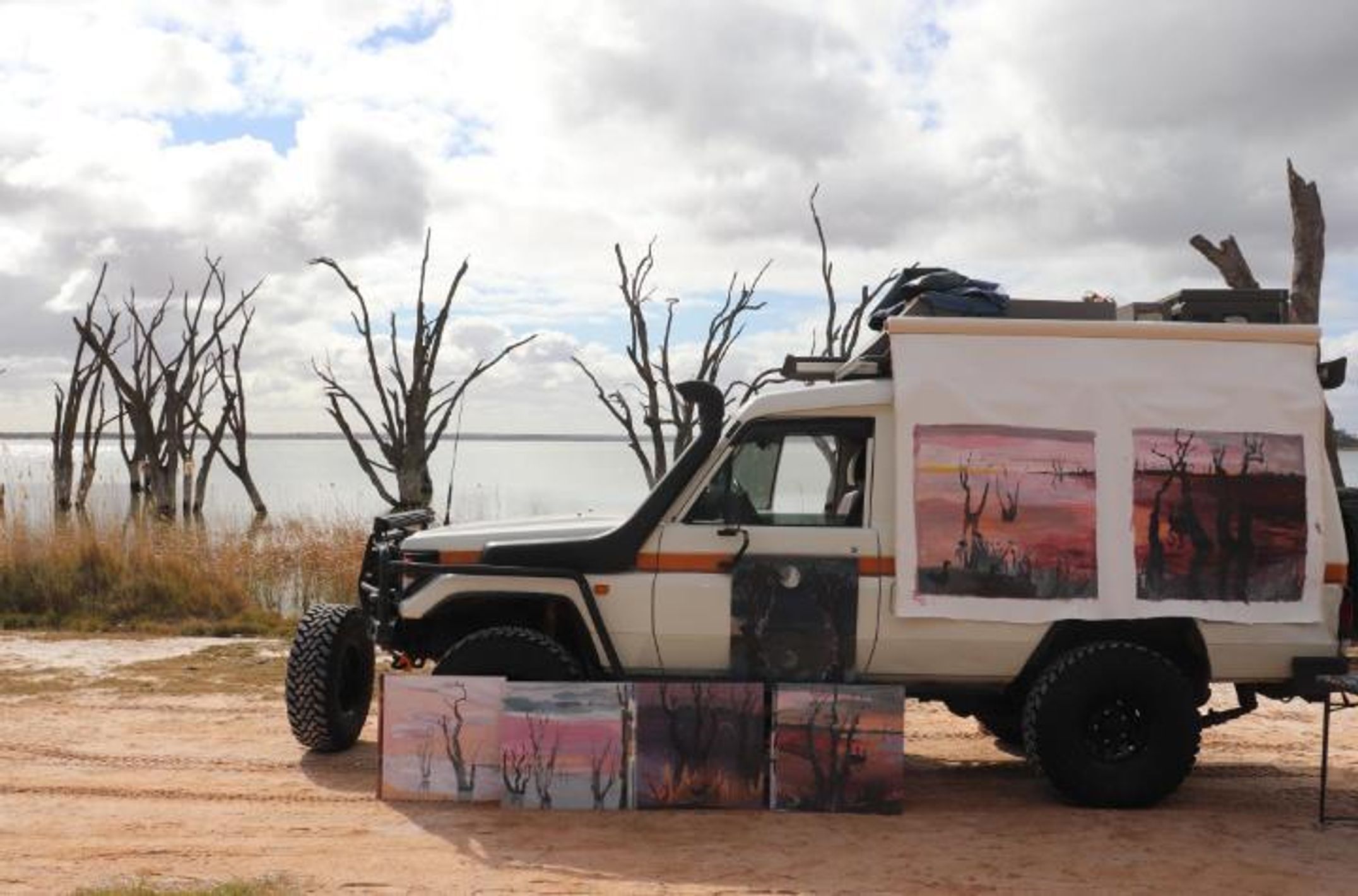 Four wheel drive vehicle with several artworks leaning on it parked beside a lake