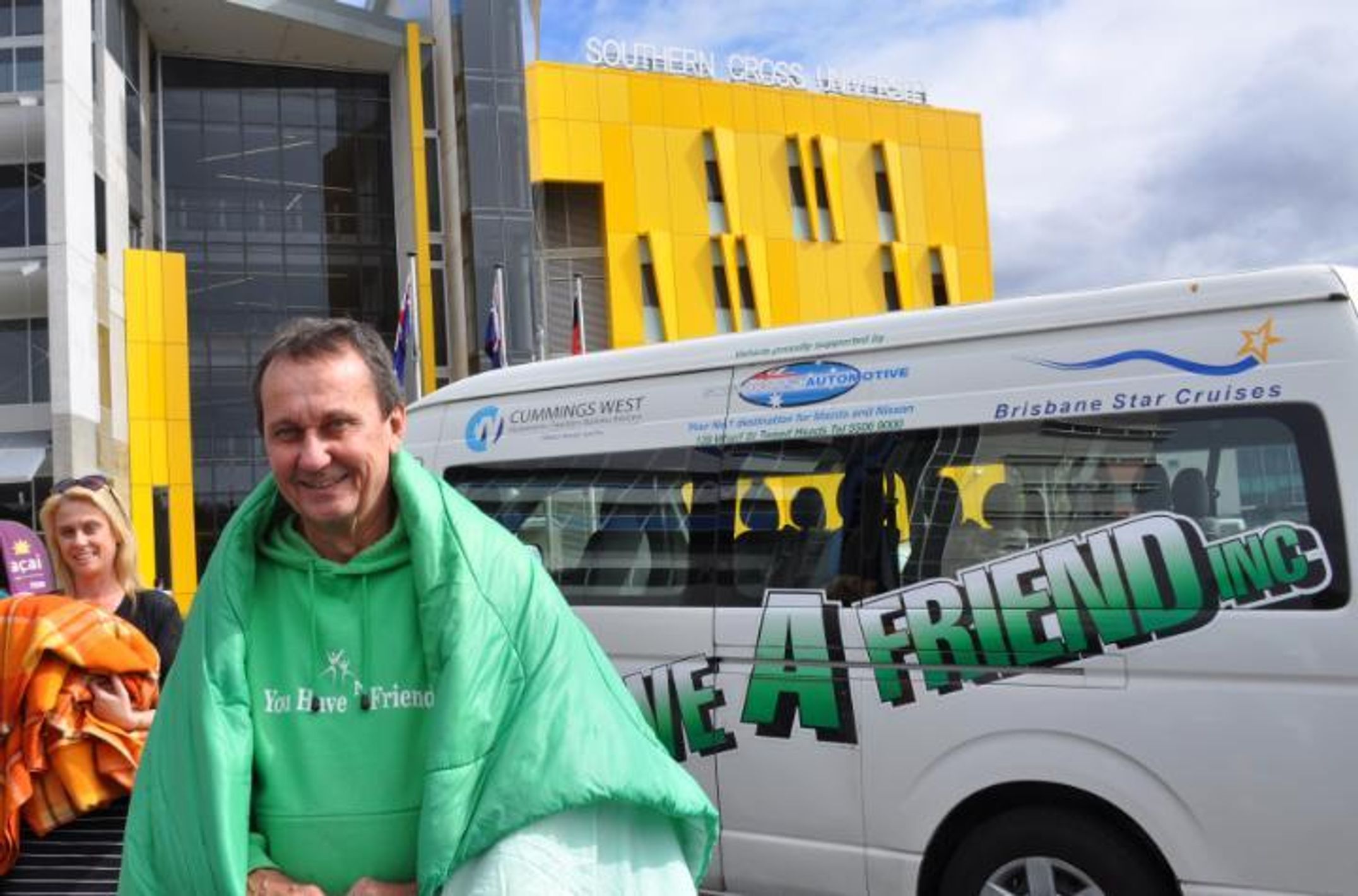 Man wrapped in a green sleeping bag standing in front of a van and a University building