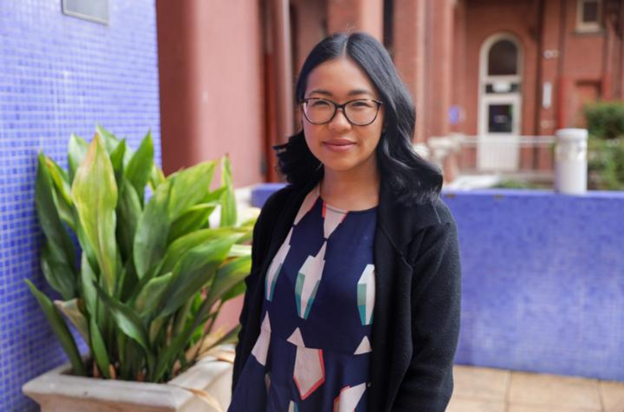 A woman with dark hair and spectacles standing in front of a building