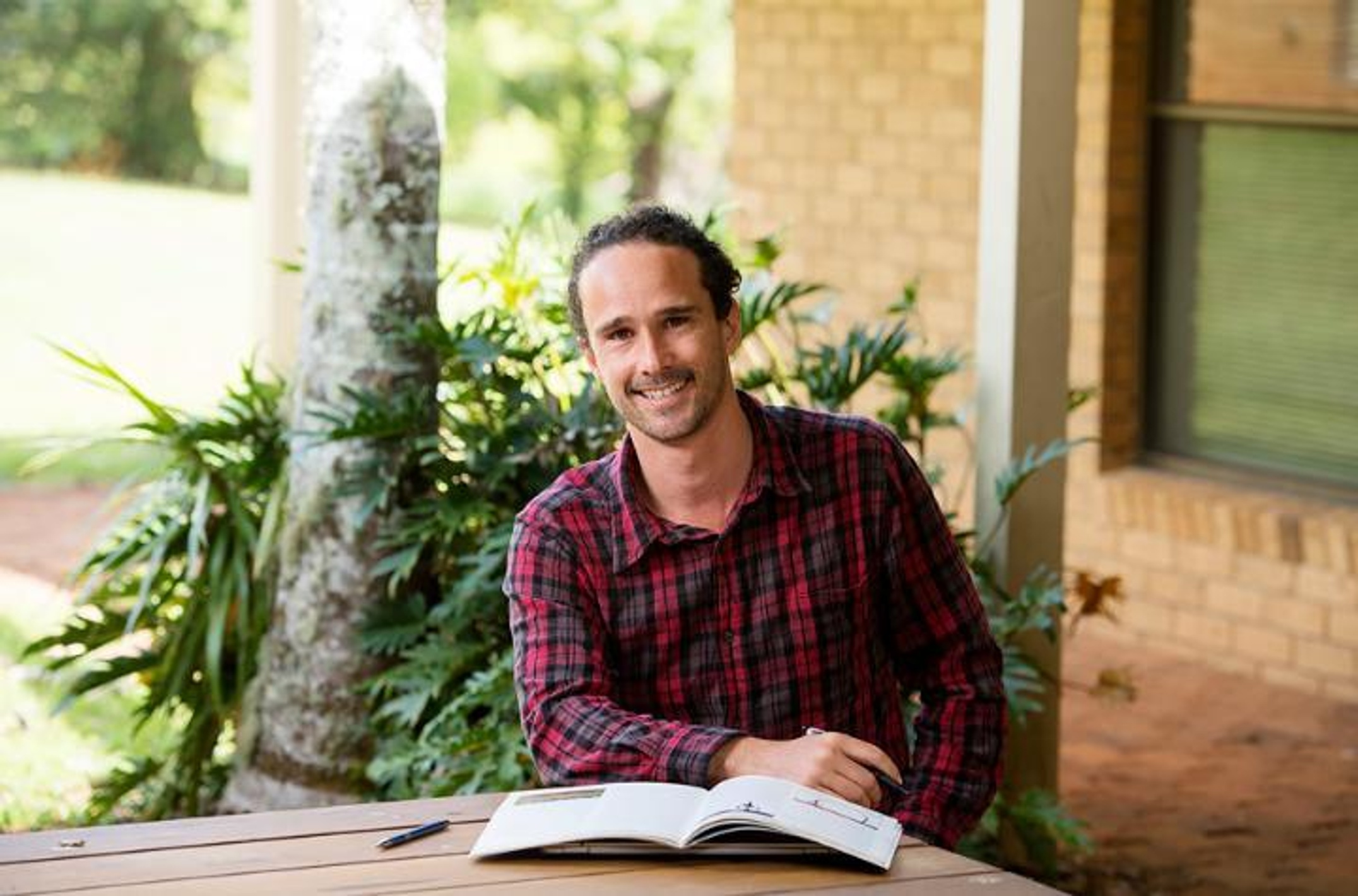 A man with a beard in a red check shirt sitting at a table with a textbook