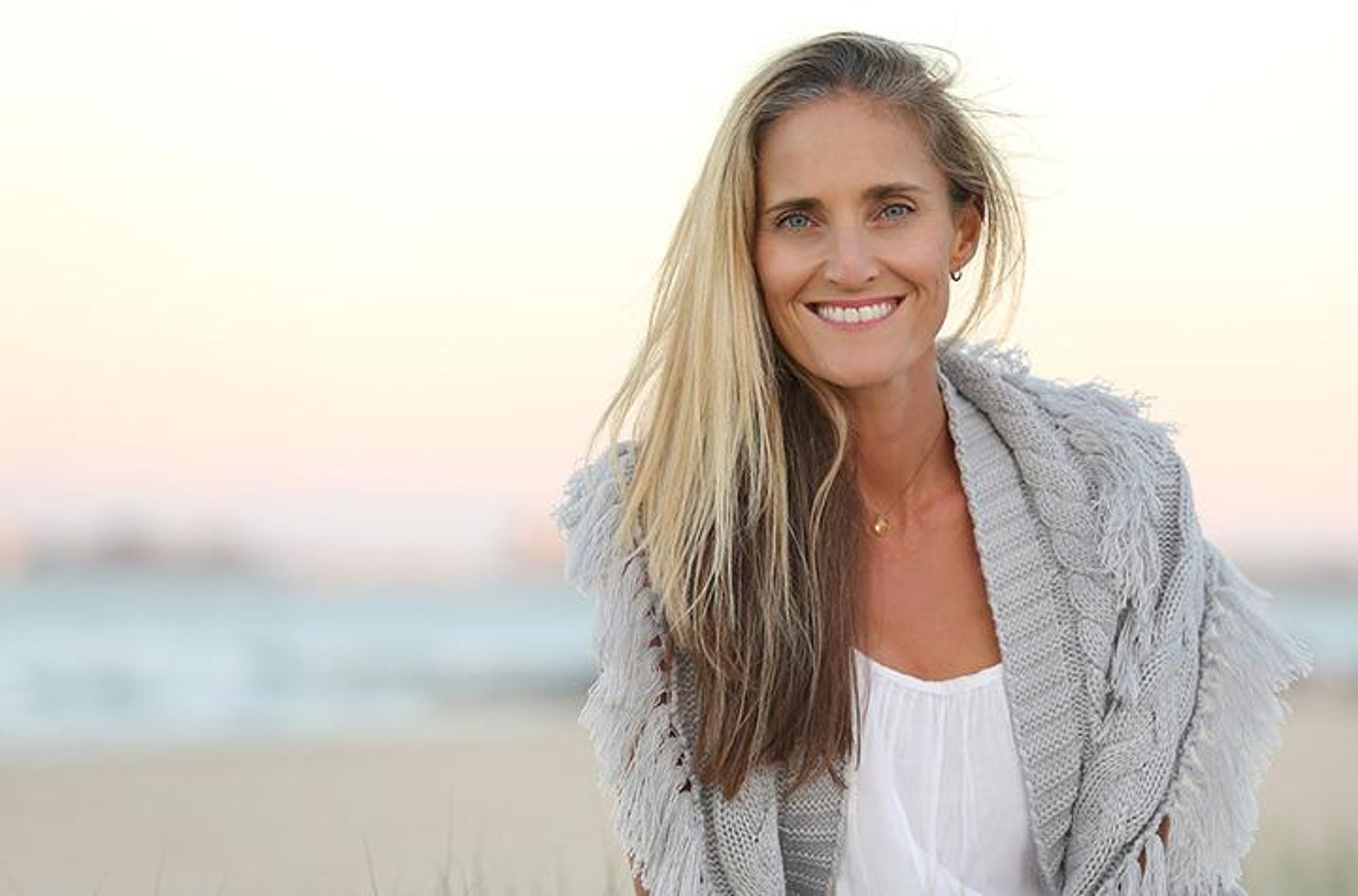 A woman with long blonde hair wearing grey jumper at the beach