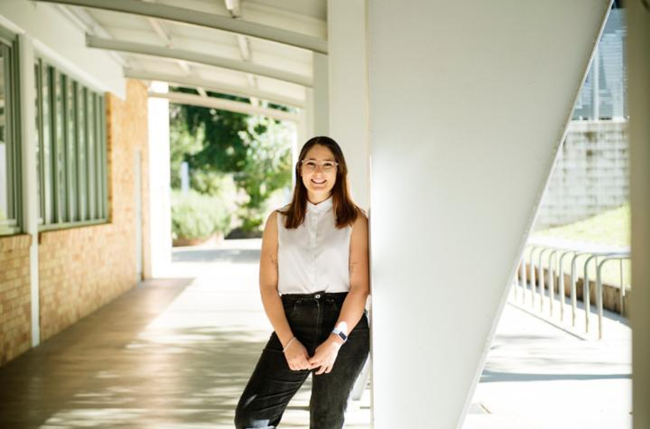 Woman in white shirt and black jeans outside leaning against a building structure