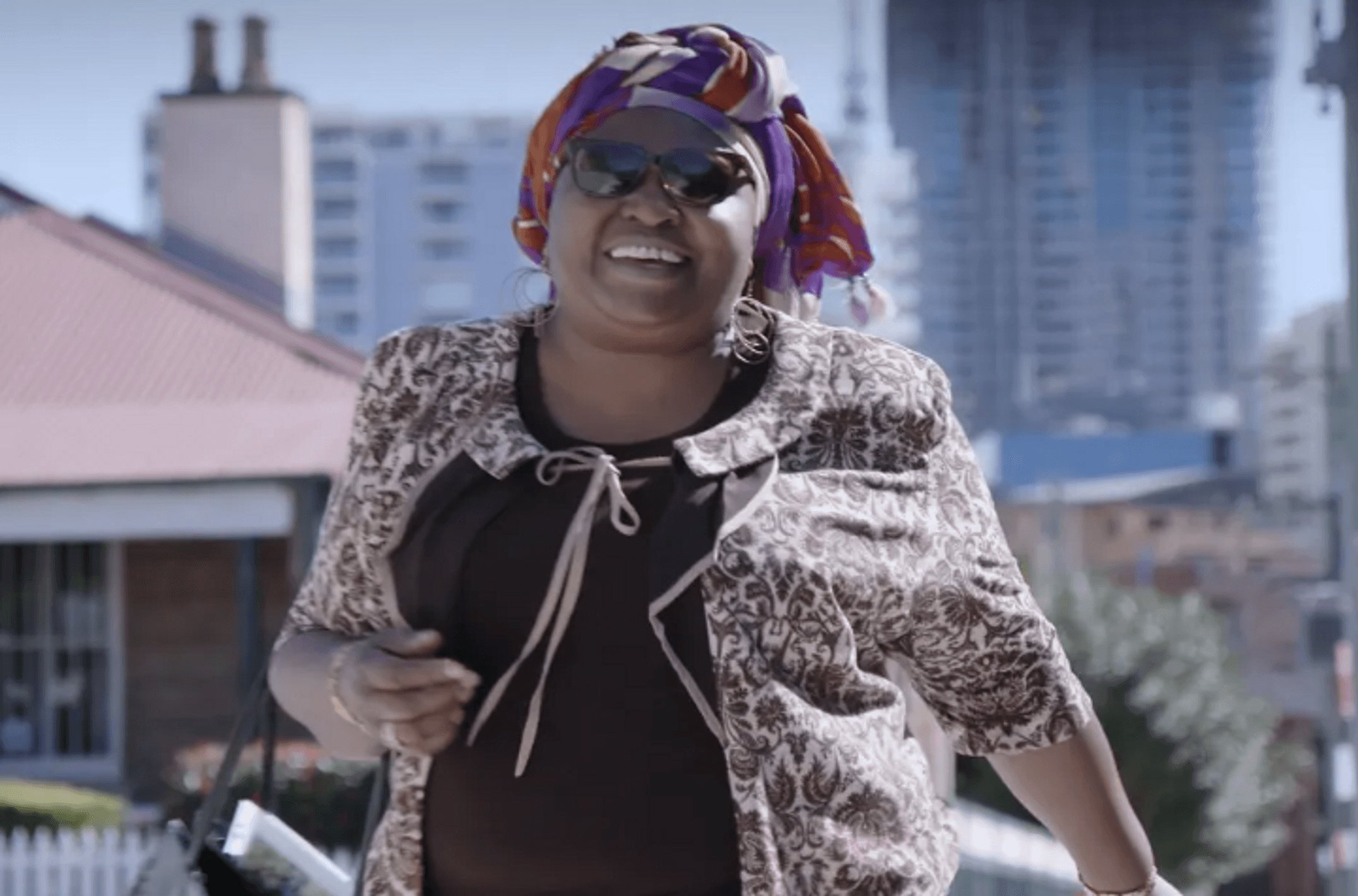 A smiling woman wearing sunglasses, handbag over arm, walking in a city