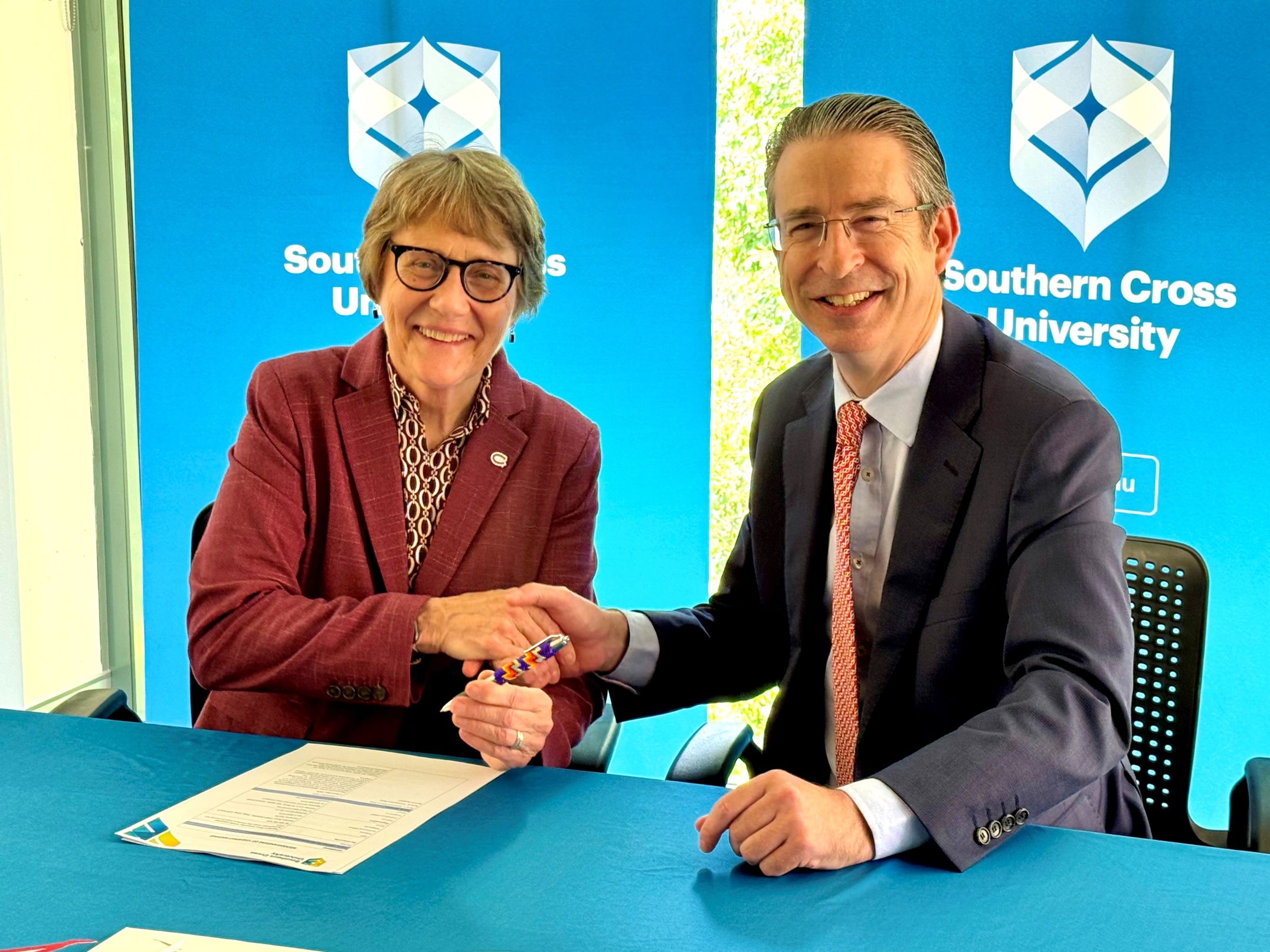 Two people shake hands at a desk, a paper agreement on the table  in front of them