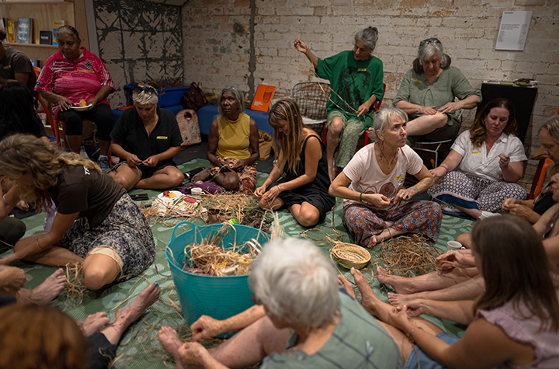 A group of Indigenous and non-Indigenous women sitting on the floor making string from plant fibres.