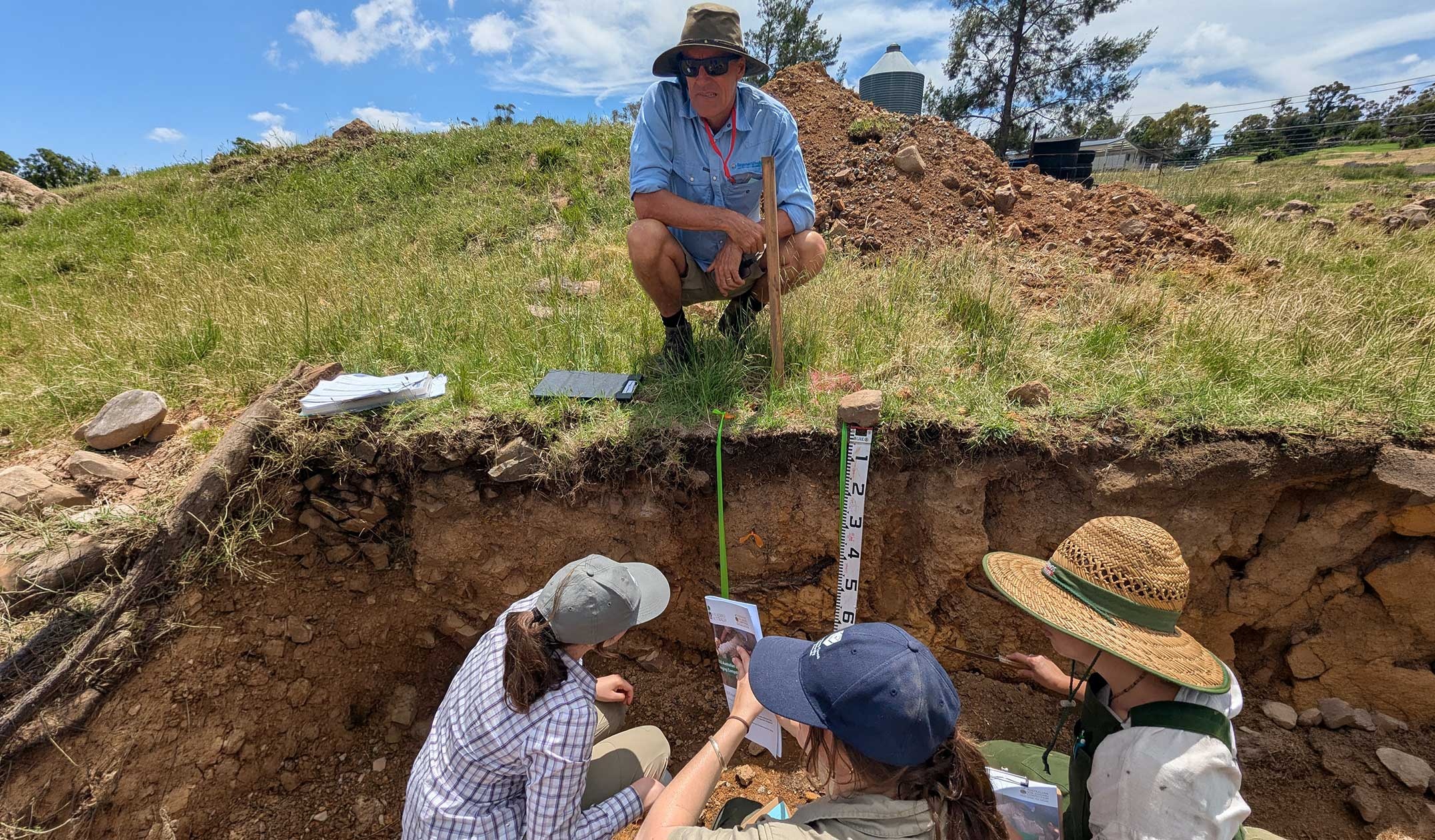 Three people in a soil pit measuring and one man standing above pit