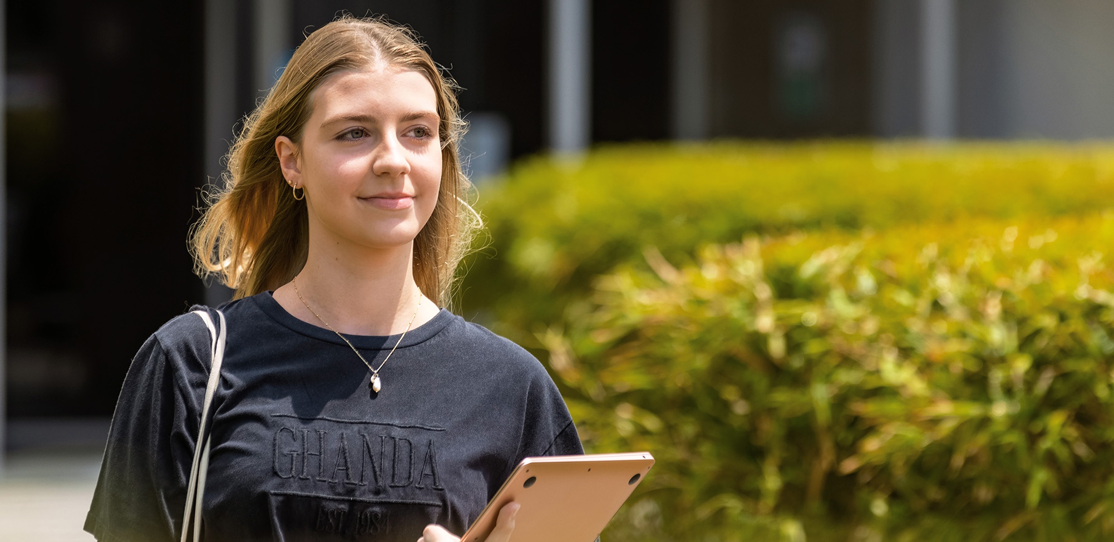 A student walks outside holding a laptop and smiling at the Southern Cross University Gold Coast campus