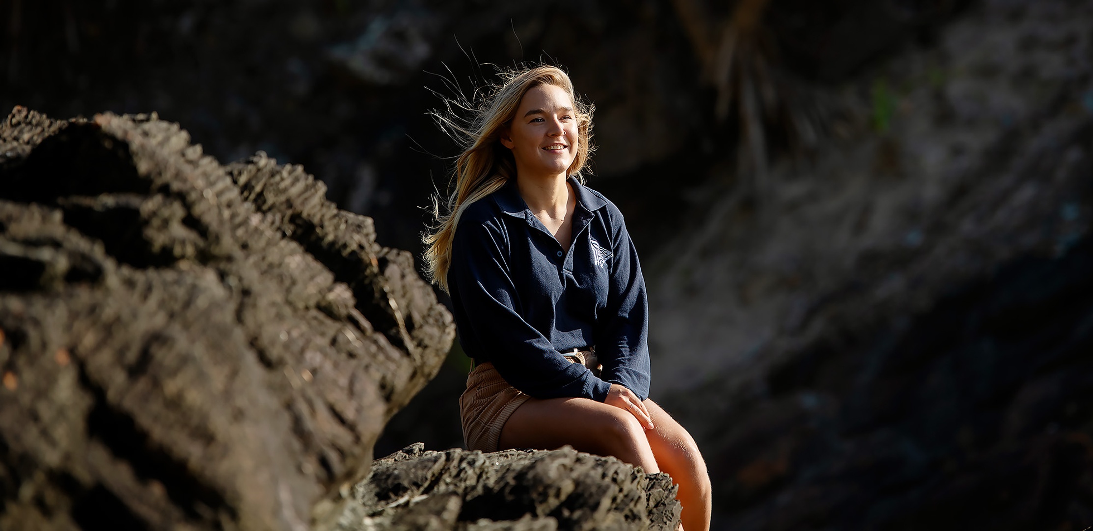 A marine science alumni sits in a rocky coastal landscape, she is wearing a blue long sleeved shirt and brown shorts and is smiling off to the side of the camera