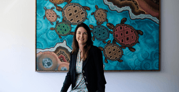 Doctor of Indigenous Philosophies graduate Kylie Day sitting in front of painting