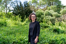 A woman faces the camera, greenery in the background