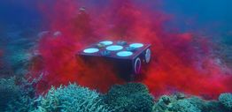 A clear box releasing a red dye, placed on the ocean floor surrounded by coral reef