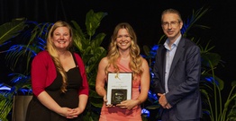 Two women and man smiling at camera. Woman in centre holding an award