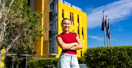 Woman in red shirt smiling with arms crossed in front of yellow building with Southern Cross University written on the roof