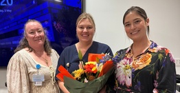 Three women smiling, the middle person holding bouquet of flowers