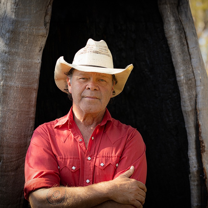 man wearing western style hat, red shirt standing in front of tree