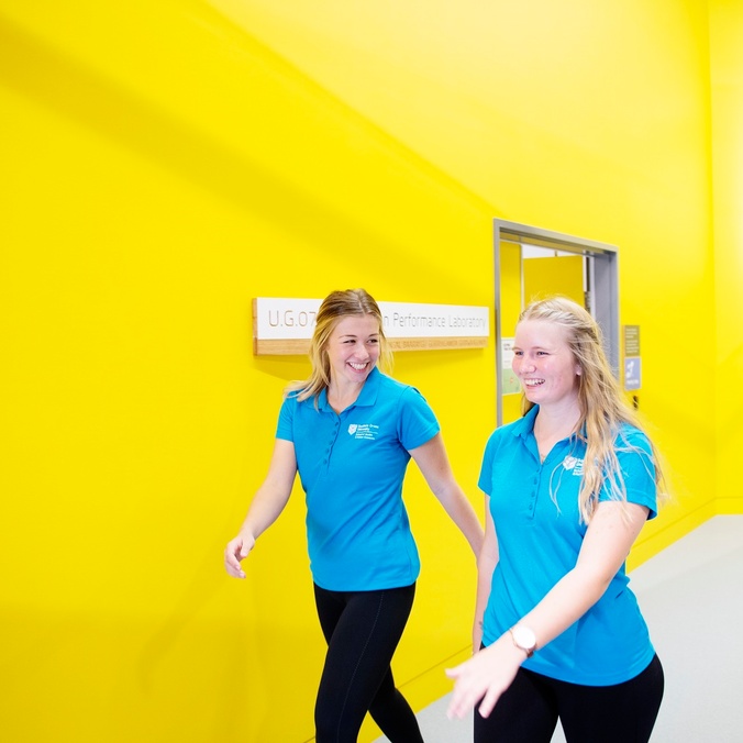 Two students in teal shirts walking past a yellow wall