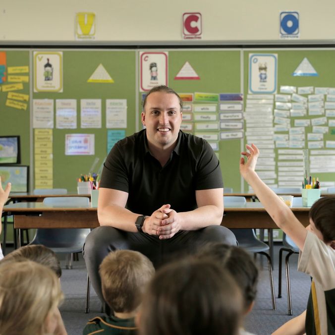A male teacher faces the camera and around him sit primary school children