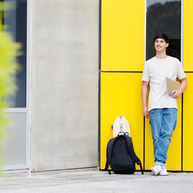 A student leaning on a building with a backpack and notebook.
