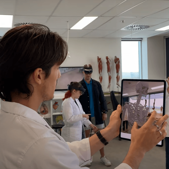 A student holds an ipad, other students wearing goggles in the background