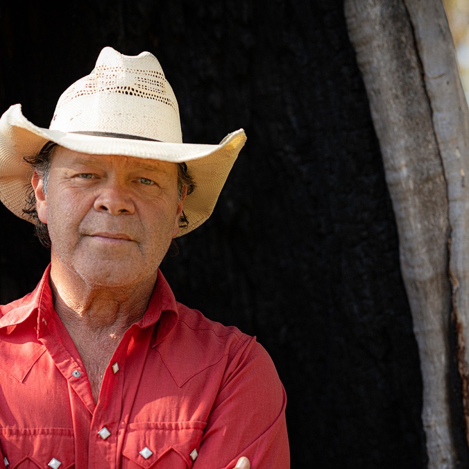 Man in cowboy hat standing in front of large tree trunk