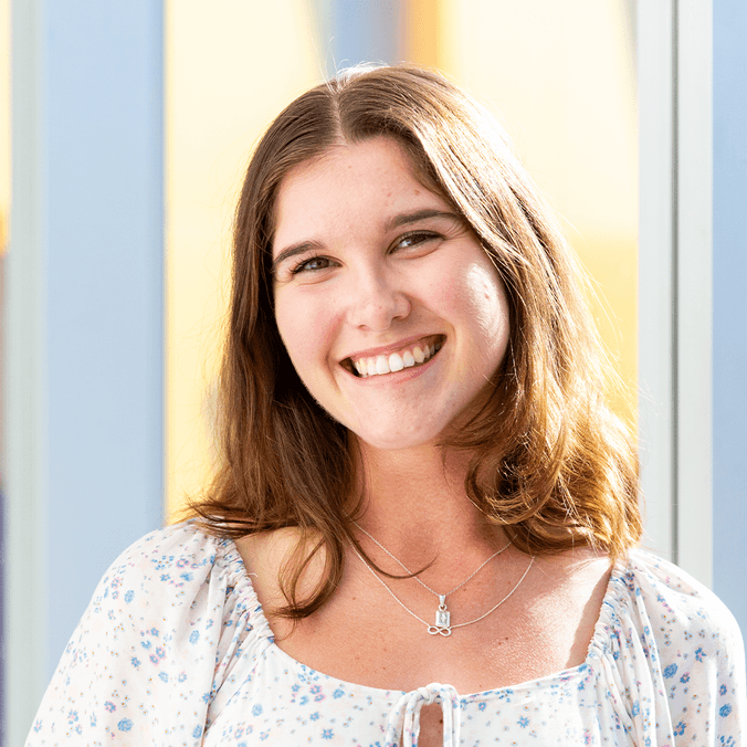 Student with brown hair in white blouse smiling with yellow building in background