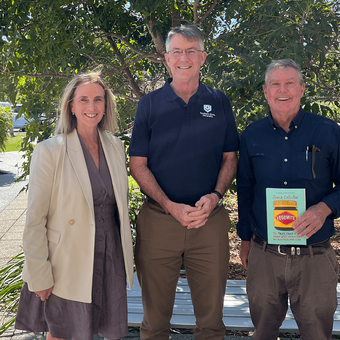 Three people posing in front of greenery, one holding a book with a jar of vegemite on the cover