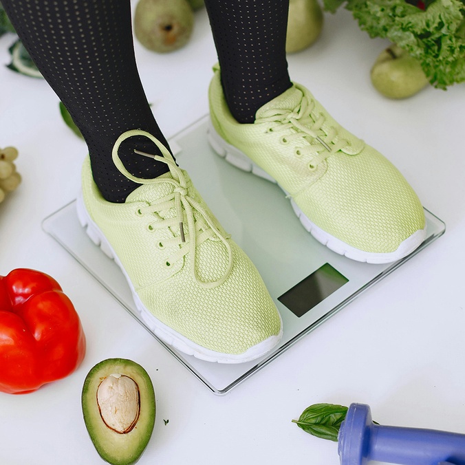 Person standing on weight scales surrounded by tape measure and fresh vegetables