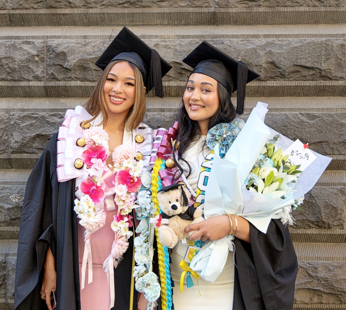 Two graduation ceremony participants wearing academic gowns and mortarboards pose in front of a stone wall. Both are wearing decorative graduation leis and holding large bouquets of flowers. One person is also holding a small teddy bear dressed in graduation attire. The image highlights celebratory graduation accessories and gifts.