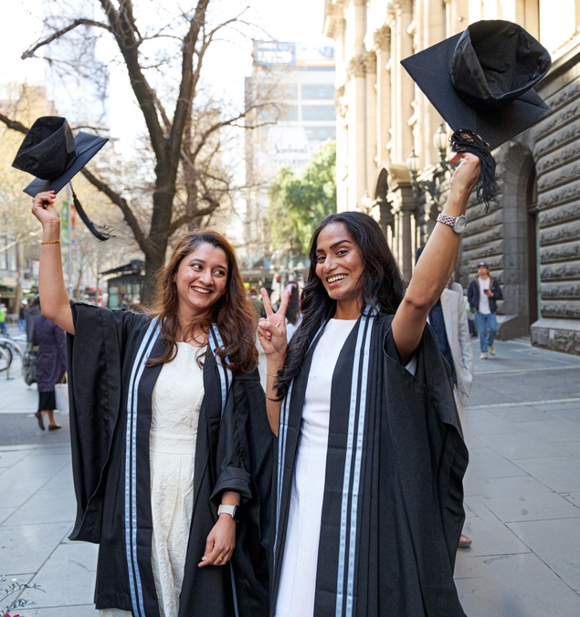 Two graduation ceremony participants wearing black academic gowns stand on a city street outside a large stone building. Each person is holding a mortarboard hat raised above their head. One person is making a peace sign with their free hand. Trees, pedestrians, and urban buildings are visible in the background.