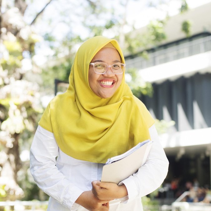 A student with a head scarf and glasses smiling and holding books