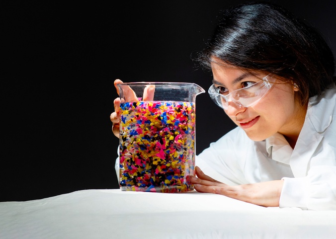 Dr Priya Borpatra Gohain BSc, MSc, PhD (SCU)  in a white lab coat looking into a large beaker or water filled with brightly colour coloured plastic particle's at Southern Cross University Lismore Campus