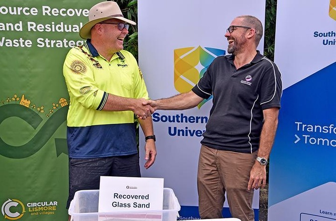 Two men shaking hands in front of a container of recycled glass