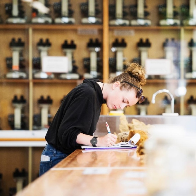 young person, leaning over bench writing on clip board, shelves of equipment behind her