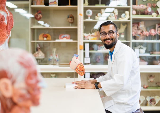 Arpan Kuwar an international student studying Nursing wearing a white lab coat and sitting in a lab looking a model of a heart at ӣֱ Lismore Campus