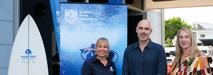 Two women and one man standing with banners and a surf board behind them