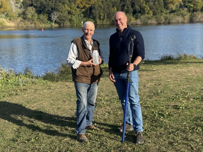 Two men stand at a river's edge holding sampling equipment