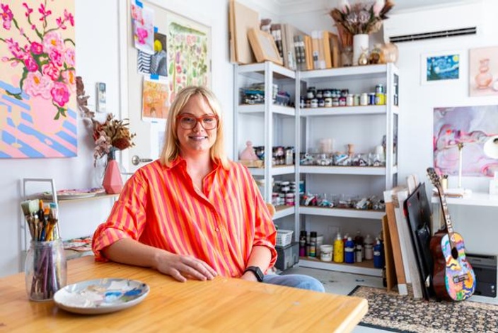 Woman in bright striped shirt sitting at desk smiling, shelves of paint in background