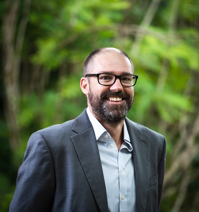 A man in a jacket and business shirt smiling at the camera against a green background