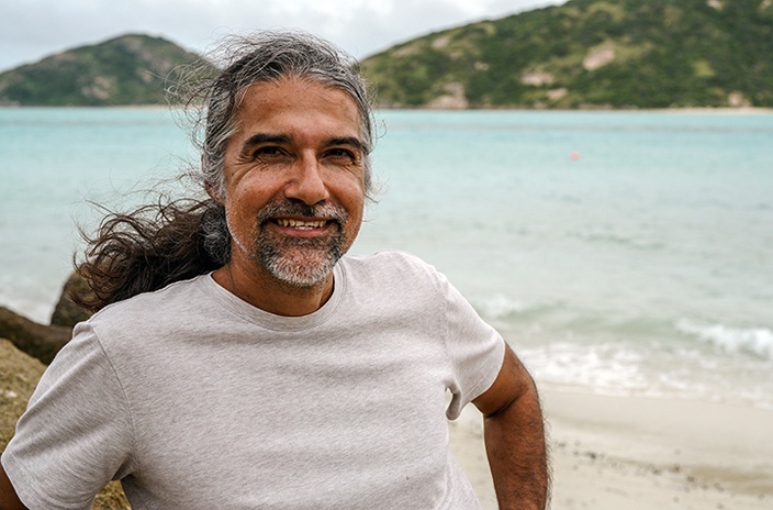 A man in a white t-shirt with the ocean and a hill in the background