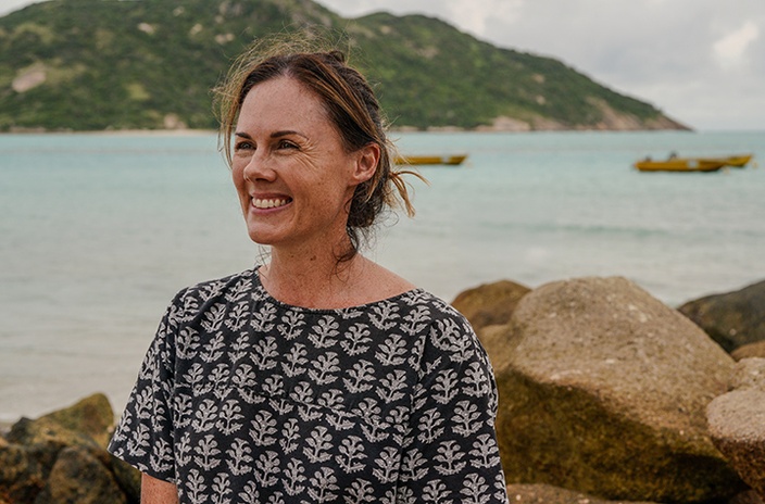 A woman in a patterned shirt with the ocean and a hill in the background