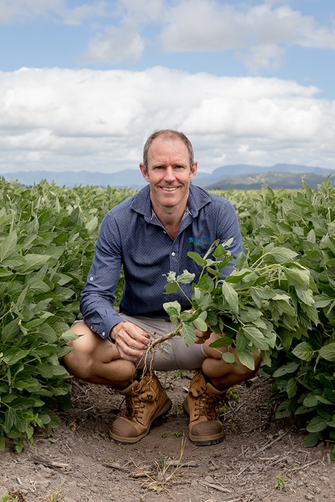 A smiling man wearing a blue shirt squatting in a paddock holding a crop in his hands