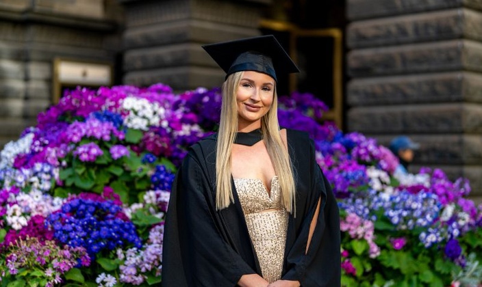 Female with blonde hair in graduation robe and cap in front of flowers
