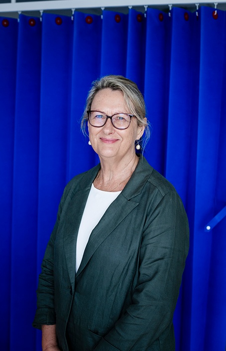 A woman wearing a green jacket and glasses in front of a blue curtain