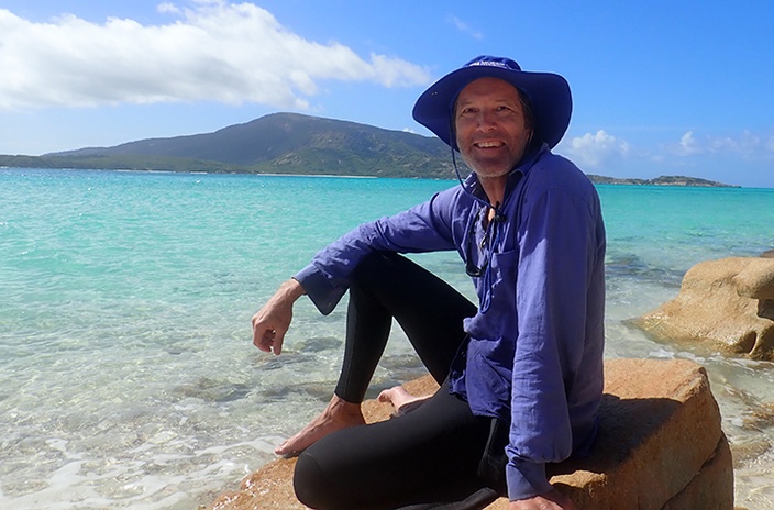 A man in a wetsuit and wearing a hat sitting on the beach