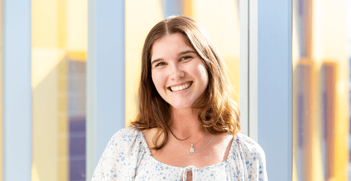 Student with brown hair in white blouse smiling with yellow building in background