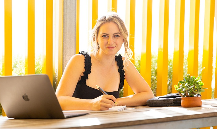 Brighley Dangerfield, SUC Business student sits at the Gold Coast campus cafe on a tall wooden table with pen, paper and laptop in front of her and bright yellow pillars behind her