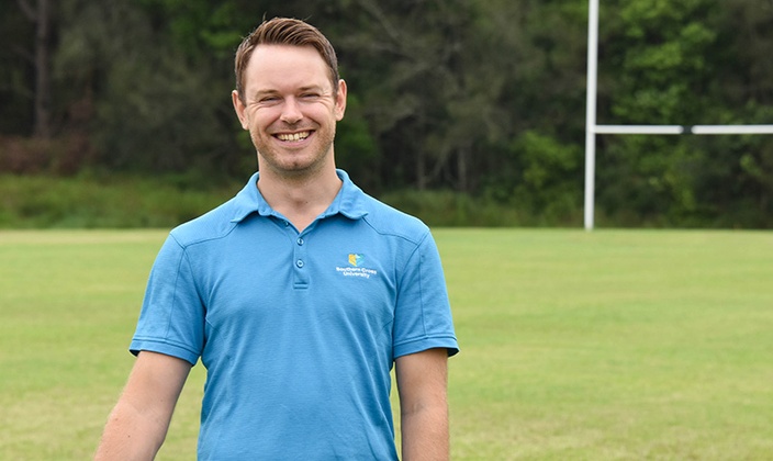 Associate Professor Christian Swann stands smiling on a rugby field with his blue Southern Cross University polo on