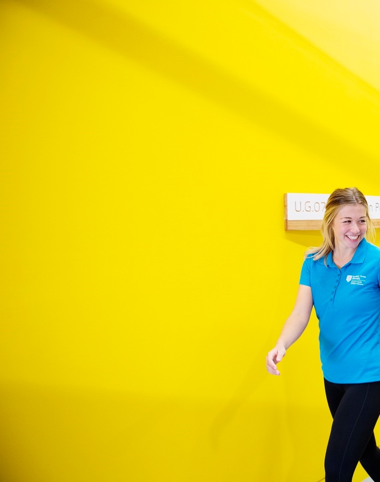 Two students in teal shirts walking past a yellow wall